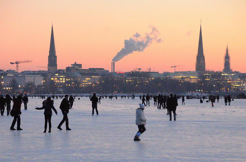 55_5950 Bis spt in den Abend geniessen die Hamburger die winterliche Atmosphre auf der zugefrorenen Alster - die untergehende Sonne frbt den Hamburger Himmel rot; aus den Schornsteinen des Heizkraftwerks in der Hafencity steigt weisser Rauch. Winter in der Hansestadt Hamburg - Sonnenuntergang an der zugefrorenen Alster.