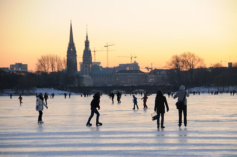 56_5925 Bis in den Abend hinein nutzen die Hamburger und Hamburgerinnen die Mglichkeit auf der zugefrorenen Auenalster Schlittschuh zu laufen oder spazieren zu gehen. Im Sonnenuntergang zeichnen sich Baukrne und der Turm des Rathauses und der Nikolaikirche im roten Abendhimmel ab. Winterliches Hamburg - Alstereisvergngen auf der Aussenalster; Sonnenuntergang.