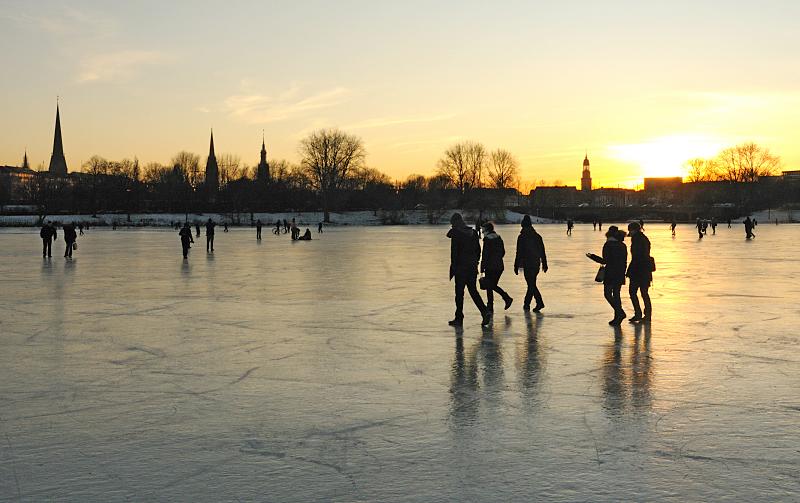59_5875 Spaziergnger dem Eis der zugefrorenen Alster gehen Richtung Hamburg - St. Georg. Das Licht der untergehenden Sonne spiegelt sich auf der blanken Eisdecke. Fotos vom winterlichen Hamburg - Abend auf der Aussenalster, Sonnenuntergang + Abendhimmel. 