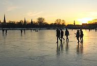 59_5875 Spaziergänger dem Eis der zugefrorenen Alster gehen Richtung Hamburg - St. Georg. Das Licht der untergehenden Sonne spiegelt sich auf der blanken Eisdecke. Fotos vom winterlichen Hamburg - Abend auf der Aussenalster, Sonnenuntergang + Abendhimmel. 