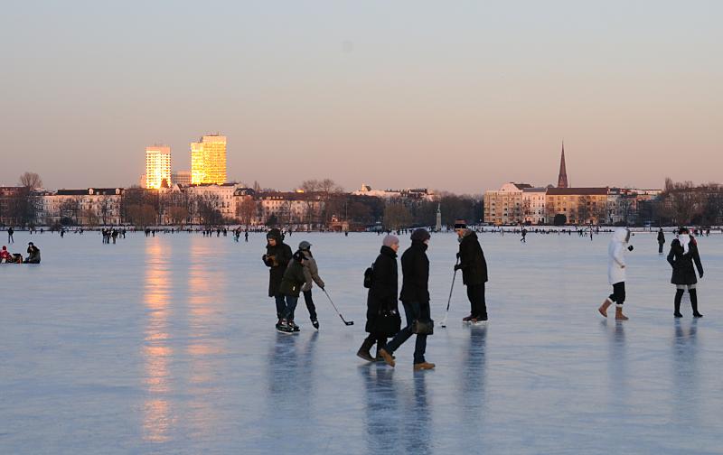 61_5885 Die untergehende Sonne strahlt die Hochhuser an der Mundsburg an - das Licht spiegelt sich auf dem Eis der zugefrorenen Alster. Zwei Schlittschuhlufer spielen Eishockey, andere gehen gegen die Klte dick mit Schal und Mtze eingemummtauf dem Alstereis spazieren. Fotos vom Winter in Hamburg - nach einer langen Frostperiode ist die Auenalster zugefroren.