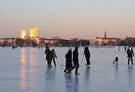 61_5885 Die untergehende Sonne strahlt die Hochhäuser an der Mundsburg an - das Licht spiegelt sich auf dem Eis der zugefrorenen Alster. Zwei Schlittschuhläufer spielen Eishockey, andere gehen gegen die Kälte dick mit Schal und Mütze eingemummt auf dem Alstereis spazieren. Fotos vom Winter in Hamburg - nach einer langen Frostperiode ist die Außenalster zugefroren.  