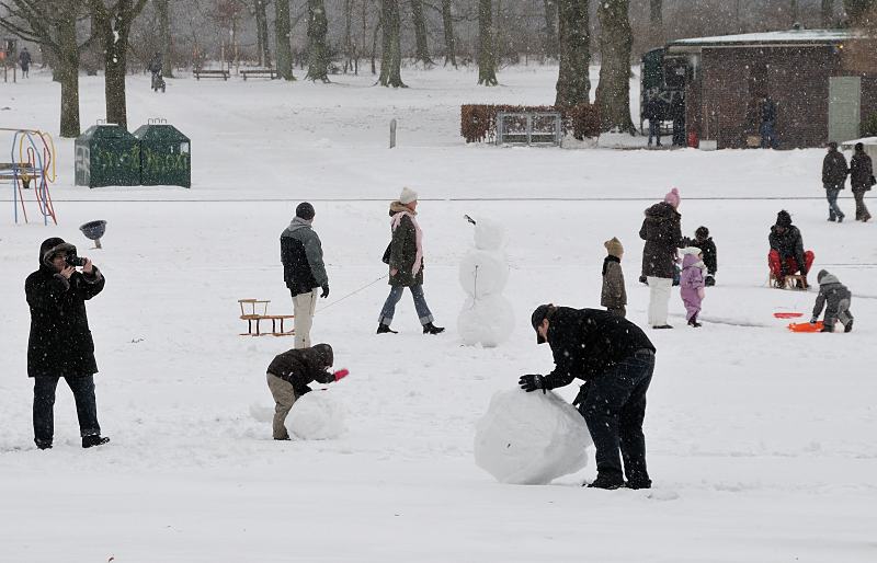 70_5017 Dichter Schnee fllt in Hamburg - im Becken des Planschbeckens, in dem im Sommer die Kinder im Wasser toben, baut ein Vater zusammen mit seinem Sohn einen groen Schneemann; die Mutter fotografiert die Aktion - andere Kinder spielen mit ihren Schlitten. Winterimpressionen von Hamburg - Vater mit Kind baut einen Schneemann.