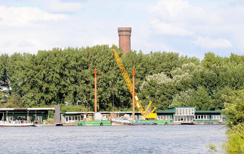 01137_8360 Blick über die Norderelbe zu den Pontons in Hamburg Entenwerder - Holzhäuser, Arbeitsbuden stehen auf den schwimmenden Anlegern, ein Schwimmkran und Barkassen / Schuten haben dort festgemacht. Hinter den Bäumen vom Elbpark Entenwerder die Spitze vom Wasserspiel / Wasserturm Hamburg Rothenburgsort.