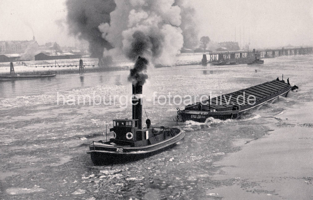 01141_33_18 Historisches Foto von der Norderelbe im Winter - Eisschollen treiben auf dem Wasser, ein Schlepper unter Dampf zieht einen Oberländer Kahn. Im Hintergrund die Zollstation Entenwerder - niedrige Lagerhäuser am Zollhafen, dahinter Wohnblocks von Hamburg Rothenburgsort.