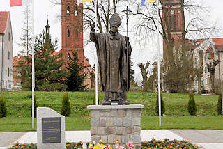 0113 Statue Papst Johannes Paul II. in Pieniężno, Polen. Im Hintergrund die Kirchtrme der evangelischen und katholischen Kirche und der restaurierte Kupferturm vom Rathaus.